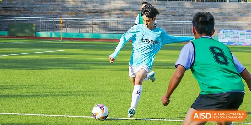 A student from the Beijing Bayi School International Department in a light blue jersey kicking a soccer/football on the green sports field.