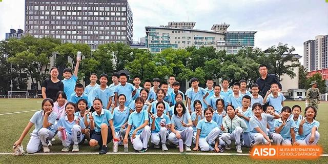 A large group of students from the Beijing Bayi School International Department, many wearing light blue shirts, kneeling and posing for a group photo outdoors on the campus lawn.