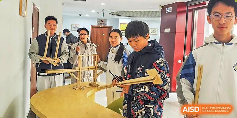 A student in a patterned jacket working on a wooden construction project with classmates looking on inside a room at the Beijing Bayi School International Department.