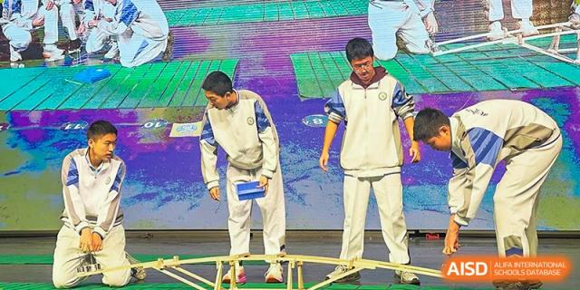 Two students from the Beijing Bayi School International Department performing a skit or demonstration on a stage, involving a wooden structure on the floor.