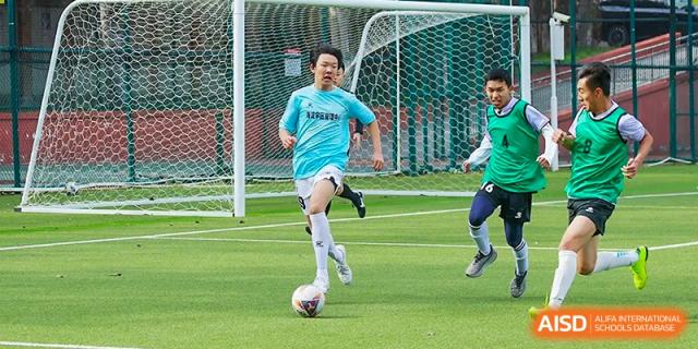 A student from the Beijing Bayi School International Department in a light blue jersey runs with the soccer/football towards the goal during a game on the school's pitch.