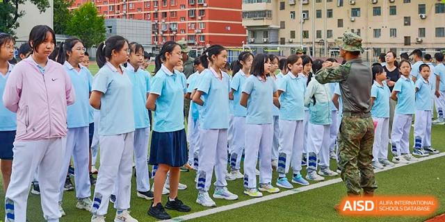 Students in light blue and white uniforms standing in a neat line outdoors on a sports surface at the Beijing Bayi School International Department campus.