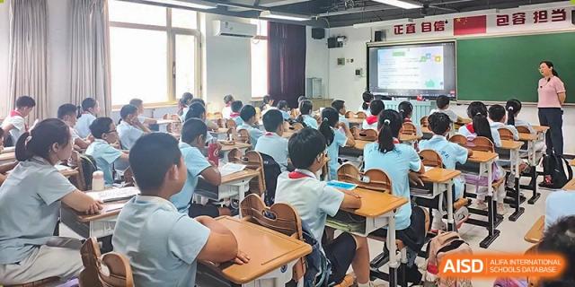 A wide shot of a busy classroom of students seated at desks, facing a projector screen at the Beijing Bayi School International Department.