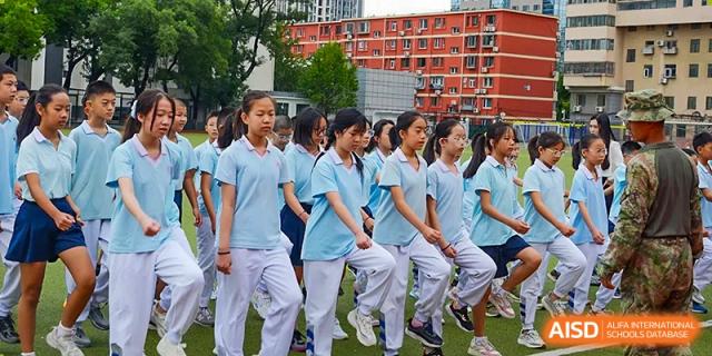 A group of female students in light blue shirts and white pants walking or running in formation outdoors near the red brick school buildings of the Beijing Bayi School International Department.