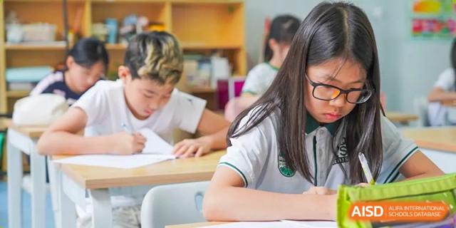 A close-up of a student in a white polo shirt writing at their desk in a bright classroom at Shanghai Hongqiao International School.