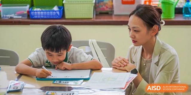 A student focused on writing, with a teacher assisting them with their desk work at Shanghai Hongqiao International School.