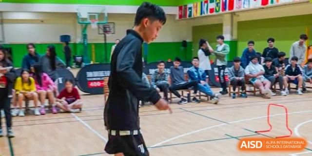 A male student playing basketball on an indoor court, with other students relaxing on the sidelines, at Shanghai Hongqiao International School.
