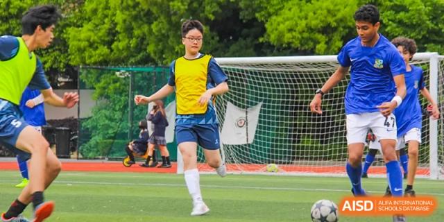 A student in a yellow vest is playing soccer on a green field and running towards the goal at Shanghai Hongqiao International School.