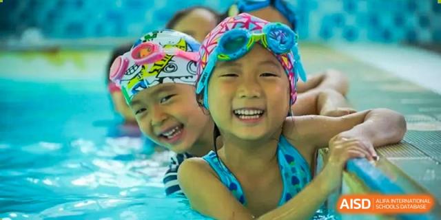 Two young, happy children wearing swim caps are smiling in a swimming pool at Shanghai Hongqiao International School.