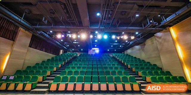 A wide shot of a large auditorium or theatre with rows of empty green seats and stage lighting set up for a performance at Shanghai Hongqiao International School.