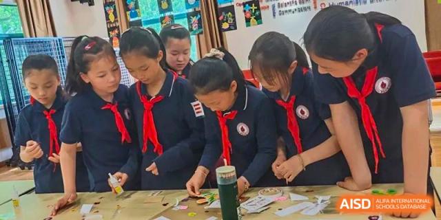 Female students from Shanghai Liaoyuan Bilingual School wearing red neckerchiefs, working on a detailed, hands-on craft or small project.