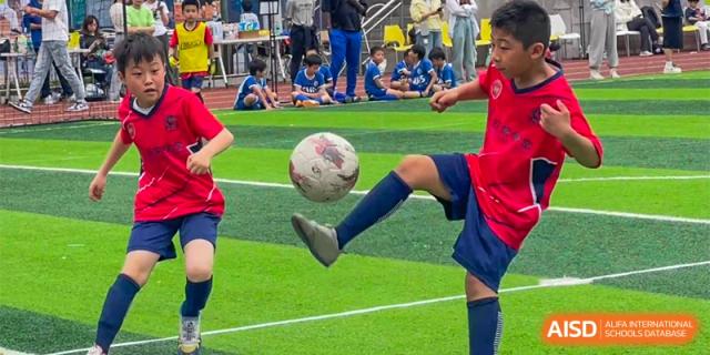 Shanghai Liaoyuan Bilingual School student in red and blue uniform playing soccer on a green turf field, kicking the ball up with their leg.