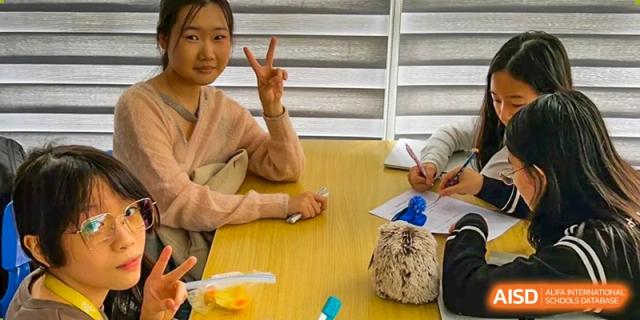 An adult giving a peace sign while sitting with children at a table, possibly during a parent/student event at QSI International School of Shenzhen.