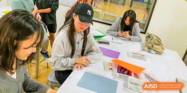 A female student from QSI International School of Shenzhen wearing a baseball cap, concentrating on writing or drawing in a notebook.