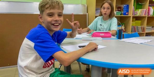 Two happy students from QSI International School of Shenzhen giving peace signs while sitting at a table in a classroom.