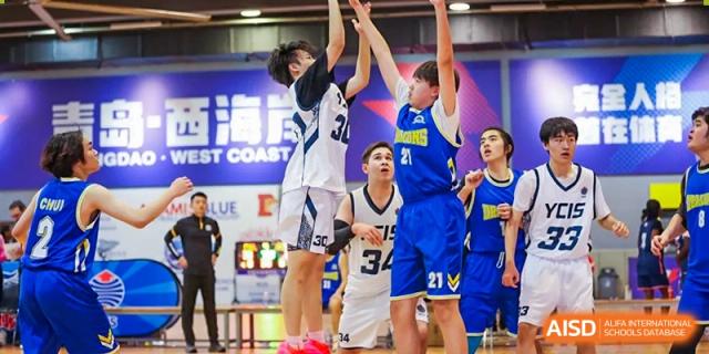 QSI International School of Shenzhen student athletes playing an intense basketball game, with players jumping to block a shot near the hoop.