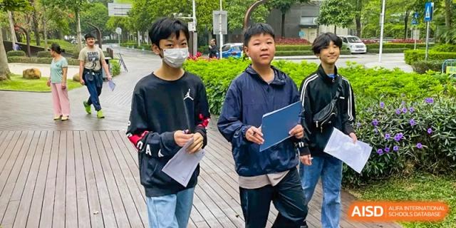 QSI International School of Shenzhen students standing outside, dressed casually and holding notebooks or folders, likely discussing schoolwork.
