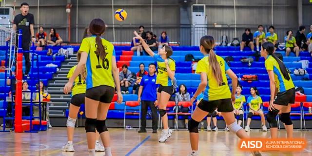QSI International School of Shenzhen student athletes playing an indoor volleyball match, with one player jumping to hit the ball over the net.