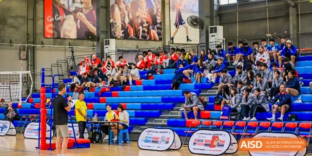 A diverse crowd of students and supporters sitting in the blue and red bleachers during a sports competition or tournament hosted by QSI International School of Shenzhen.