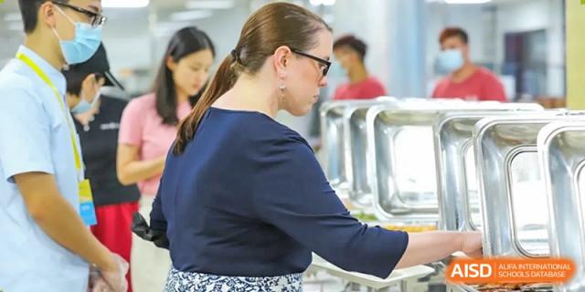 An adult, possibly a staff member at QSI International School of Shenzhen, serving food from chafing dishes at a buffet line.