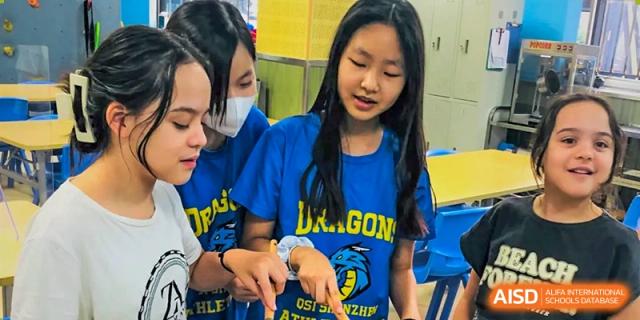 Female students from QSI International School of Shenzhen wearing blue 'Dragon' team shirts, interacting happily during an event.
