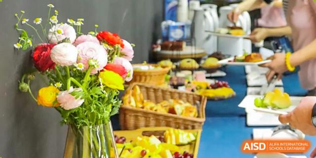 A vibrant spread of fresh fruit, flowers, and snacks displayed in baskets and serving dishes, likely for a school reception or event at QSI International School of Shenzhen.