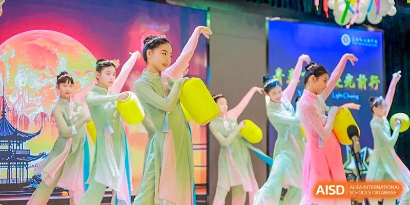Students performing a traditional Chinese dance, wearing light green costumes and holding yellow props on a brightly lit stage at Suzhou Foreign Language School.