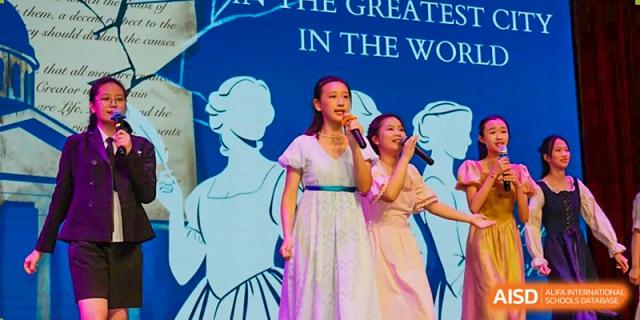 Students singing in light-colored dresses on a stage in front of a banner about achieving 'THE GREATEST' results at Suzhou Foreign Language School.