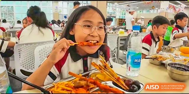 A student smiling while eating a piece of fried chicken or similar lunch meal in the school cafeteria at Suzhou Foreign Language School.