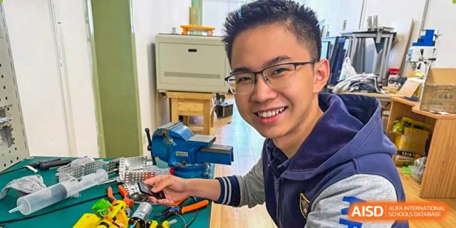 A male student smiling and working on a practical construction or technical project using a vise and tools in a workshop at Tianjin Farragut School.