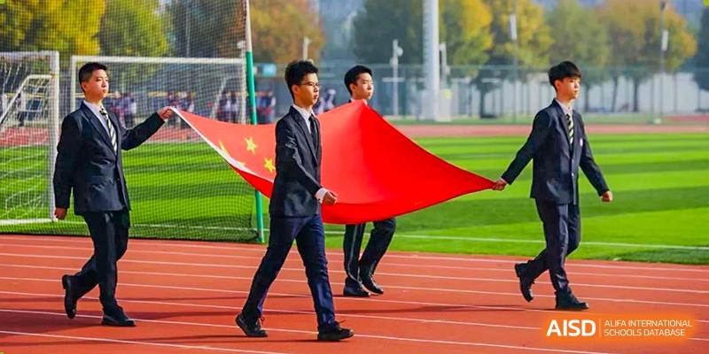 Two male students in suits walking and holding a large Chinese flag on a running track during a formal event or ceremony at Tianjin Farragut School.