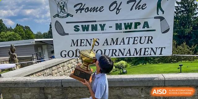A student proudly holding a large trophy near a banner advertising a men's amateur golf tournament, showcasing a student's achievement at Tianjin Farragut School.
