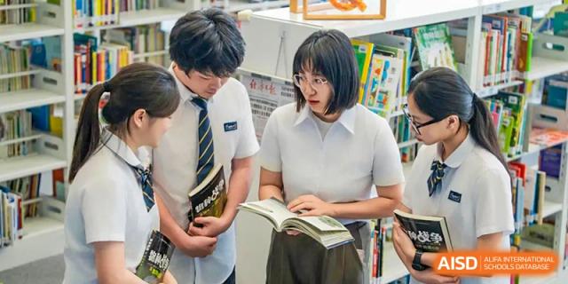 Two high school students are focused on reading books while standing amidst shelves in the modern, well-stocked library of Ardingly College Zhongshan (ACZS), promoting literacy.