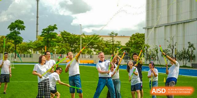 Students and staff from Ardingly College Zhongshan (ACZS) are enjoying an outdoor activity on a grassy field, making large soap bubbles with wands under a bright sky, emphasizing community spirit and fun.