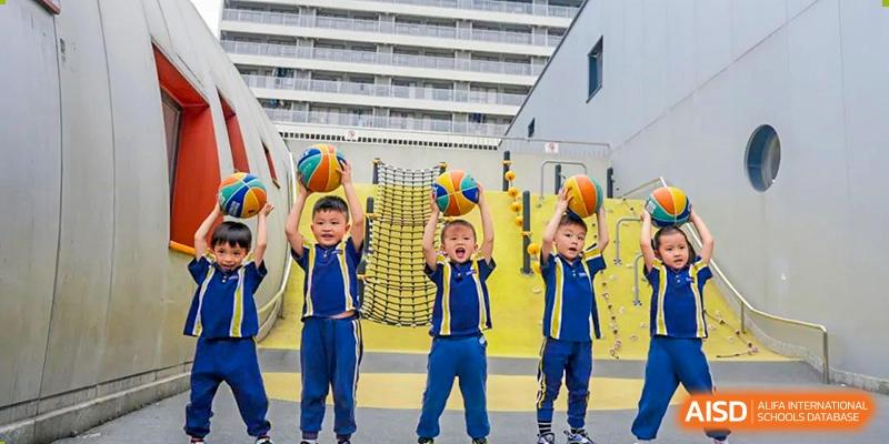 Three young, smiling children in matching blue and yellow uniforms at Ardingly College Zhongshan (ACZS) proudly hold colorful basketballs overhead on an outdoor court, highlighting early years sports and teamwork.