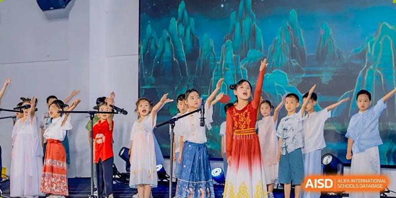 A group of children in traditional, colorful attire are singing or performing dramatically on a stage with a scenic blue backdrop at Ardingly College Zhongshan (ACZS), celebrating culture.