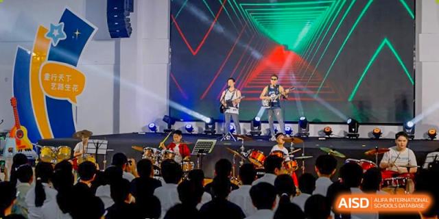 An energetic school band performs on a brightly lit stage in front of a large, engaged audience at a music event hosted by Ardingly College Zhongshan (ACZS), emphasizing performing arts.