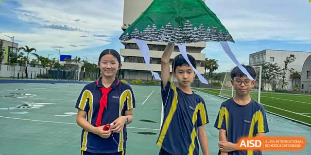 Two happy students in uniform hold up a large, kite-like creation outdoors on the field at Ardingly College Zhongshan (ACZS), promoting creative projects and outdoor activities.