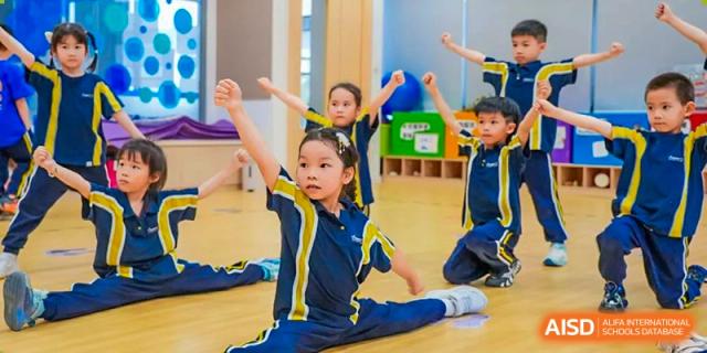 Young students in uniform are performing a synchronized dance routine or exercise, including splits, in a bright studio at Ardingly College Zhongshan (ACZS), showing flexibility and training.