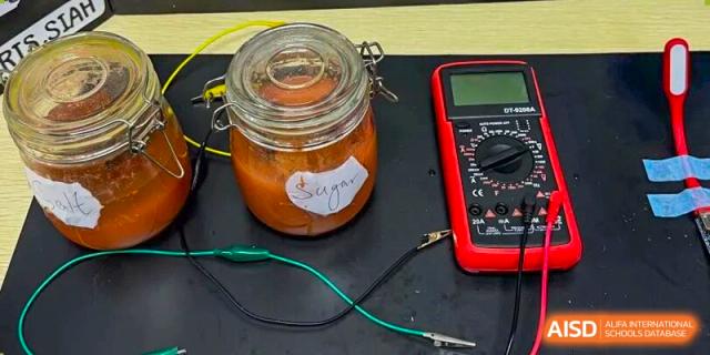 A science experiment featuring a glass jar labeled "Sugar" connected to a red digital multimeter by electrical wires at Korea International School in Shenzhen.