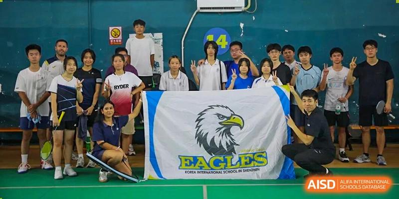 A group of students and teachers pose behind a white "Eagles" school flag on a sports court at Korea International School in Shenzhen.