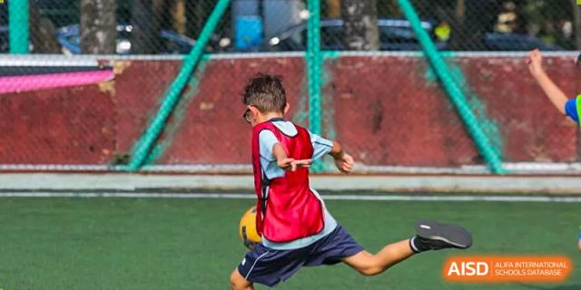 A young boy in a red mesh scrimmage vest kicks a soccer ball during an outdoor sports practice at Korea International School in Shenzhen.