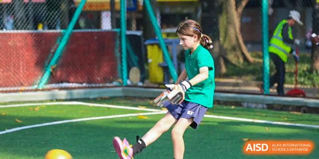 A young student in a green jersey and goalie gloves kicks a soccer ball on a lush green field at Korea International School in Shenzhen.