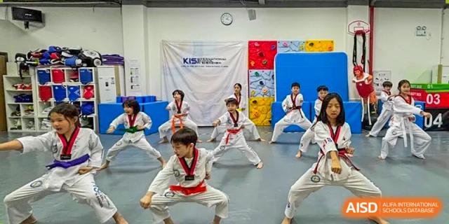 Students in martial arts uniforms practice low stances and strikes in front of a blue and yellow climbing wall at Korea International School in Shenzhen.