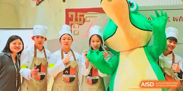Two students dressed in white chef coats and aprons pose alongside a large green mascot during a school event at BASIS International School Wuhan.
