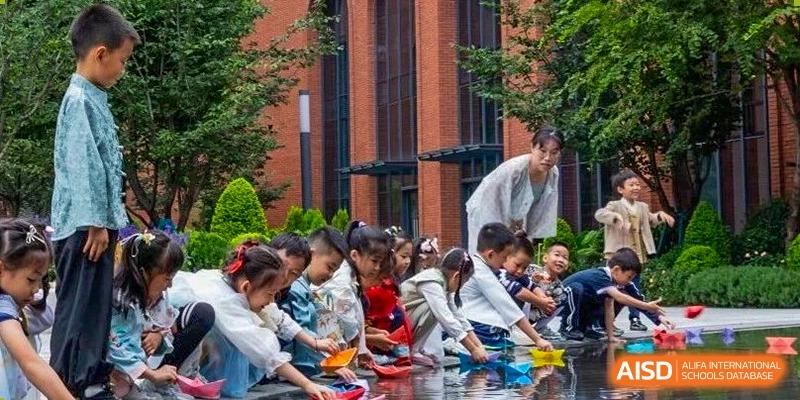 A group of young students and a teacher crouch outdoors by a water feature to sail colorful paper boats at BASIS International School Wuhan.