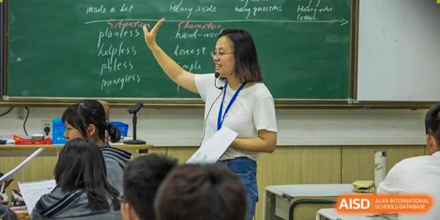 A Hangzhou Entel Foreign Language School teacher stands in front of a green chalkboard, actively leading a lively discussion with engaged students.