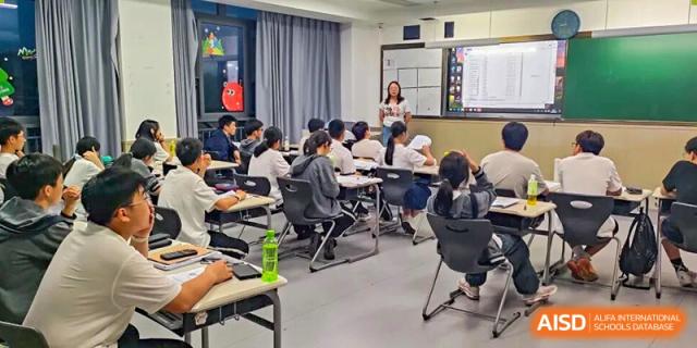 A teacher instructs students using a whiteboard in a modern classroom setting at Hangzhou Entel Foreign Language School. Students are seated at desks listening attentively.