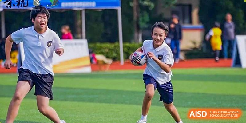 A male student with a bright smile runs across a green sports field carrying a rugby ball, actively participating in a game or drill at Hangzhou Entel Foreign Language School.