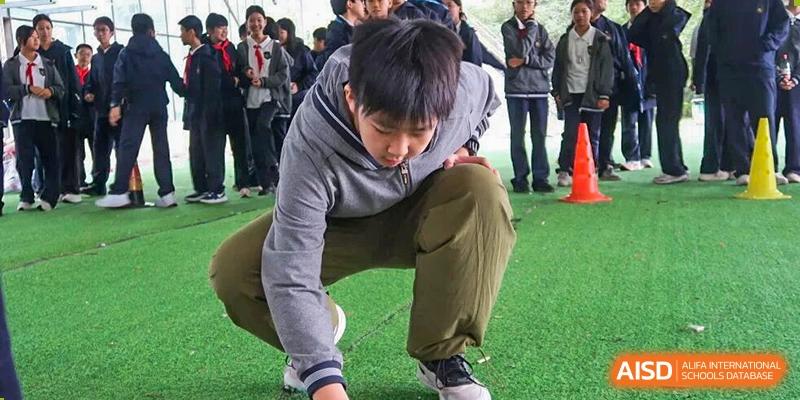 A male student crouches down on an artificial turf field, reaching out his hand, perhaps marking a spot or picking up an item, at an outdoor sports practice at Hangzhou Entel Foreign Language School.